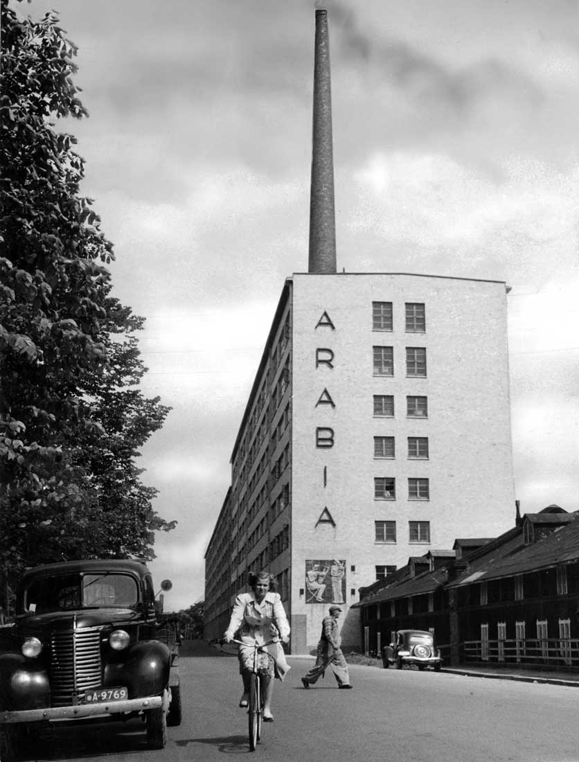 Historical black-and-white photograph from 1949 of a woman cycling past the Arabia factory building in Helsinki